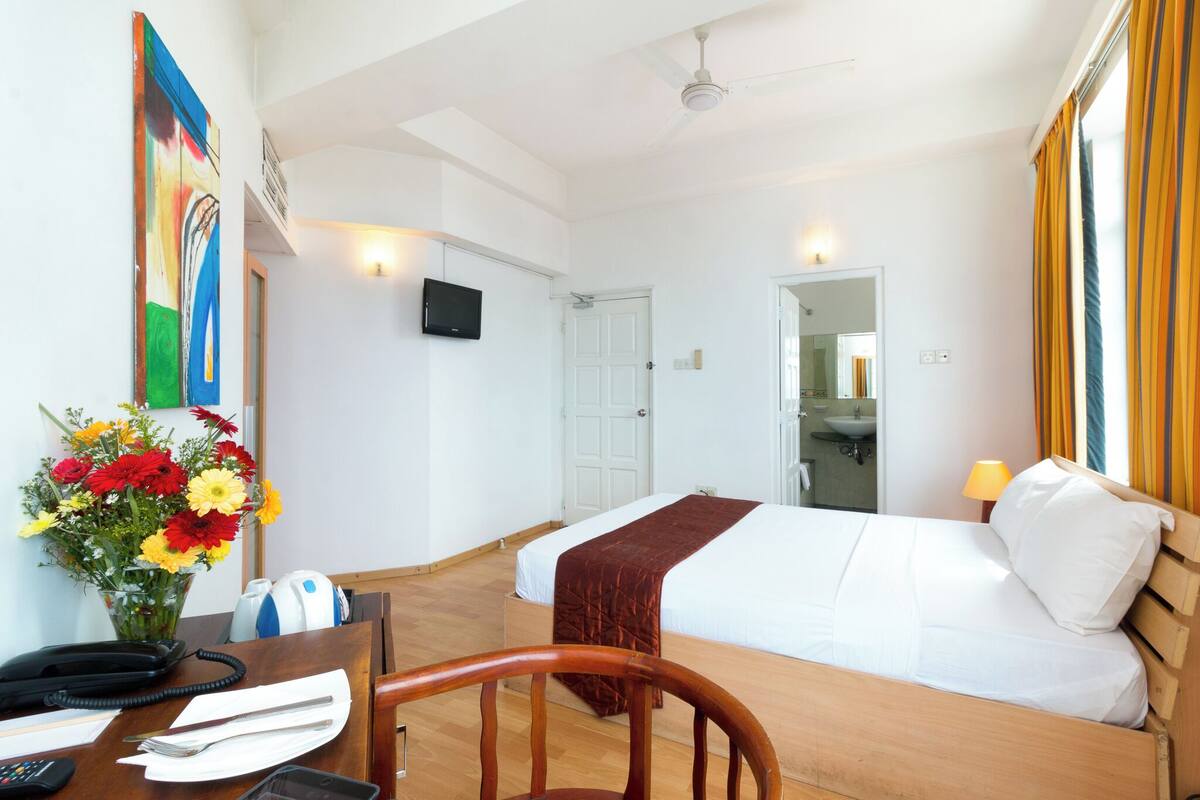 A hotel room with a bed, a desk with a telephone, a vase of flowers, and a television on the wall.
