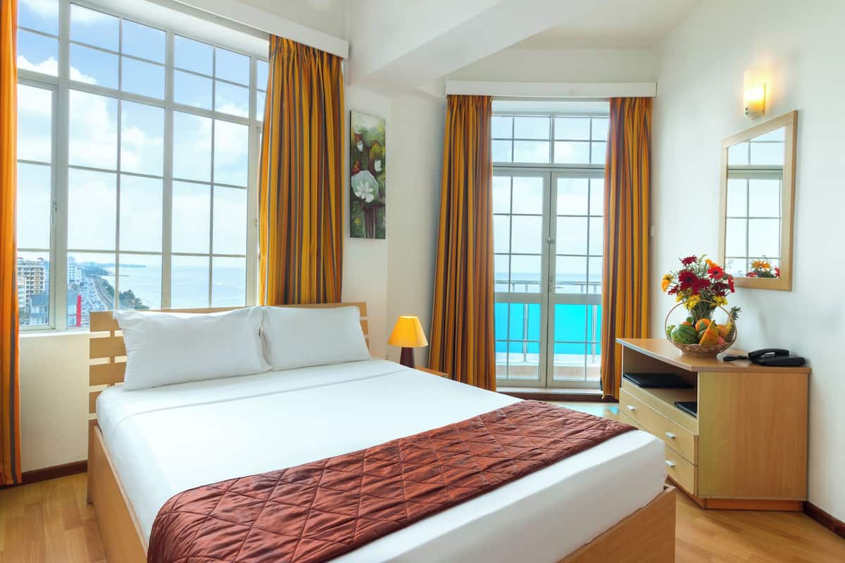 A hotel room with a large bed, a desk, a chair, a lamp, a mirror, and a view of the ocean.