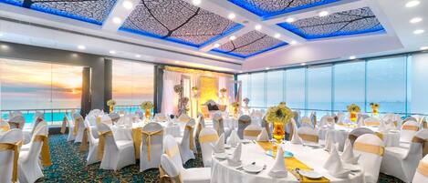 A banquet hall with round tables, white chairs with gold sashes, and a view of the ocean through large windows.