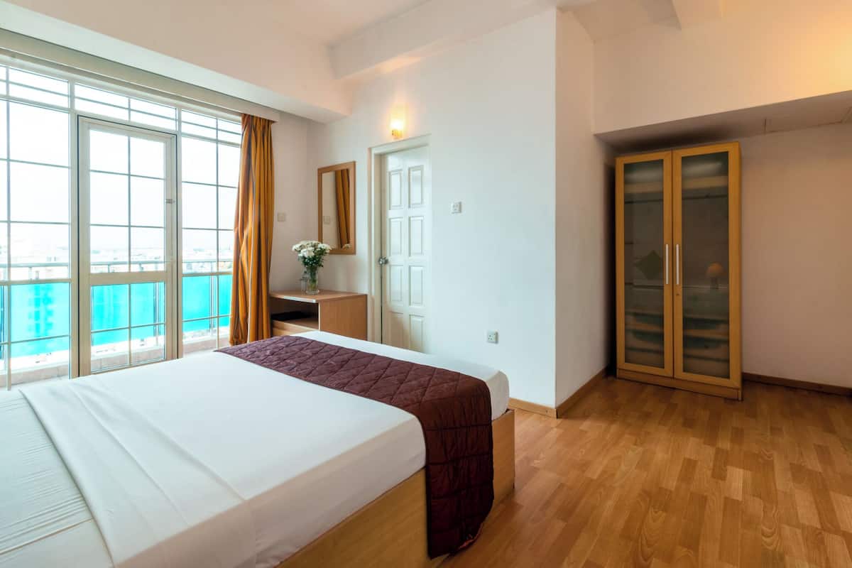 A hotel room with a large bed, a wooden floor, a balcony with a view, a small desk with a vase, and a wardrobe.