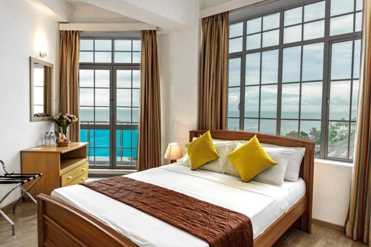 A hotel room with a large bed, a wooden desk, a chair, and a view of the ocean through large windows.