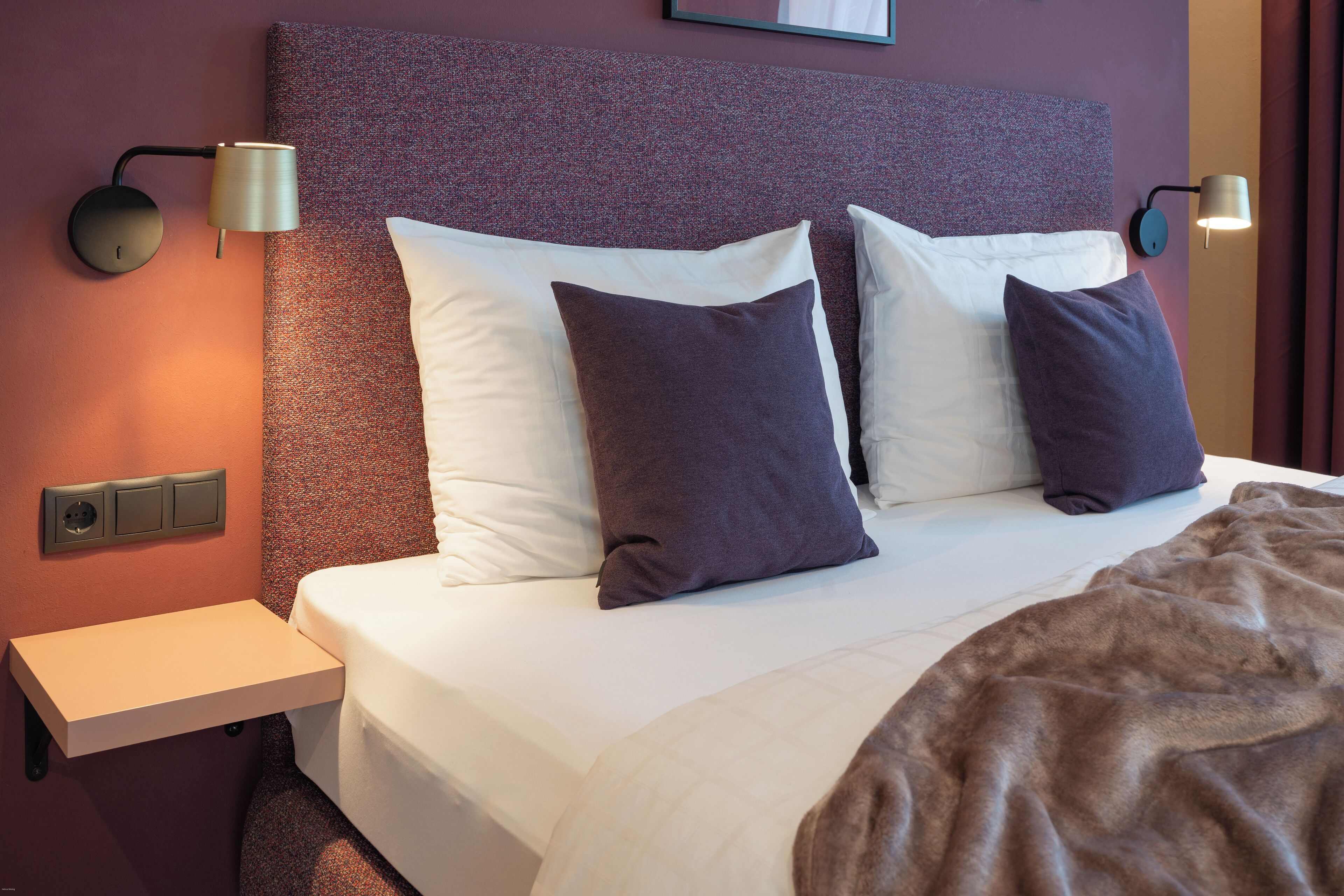 Een netjes opgemaakt bed met witte en donkere kussens, een bruin plaid en twee wandlampen aan weerszijden.