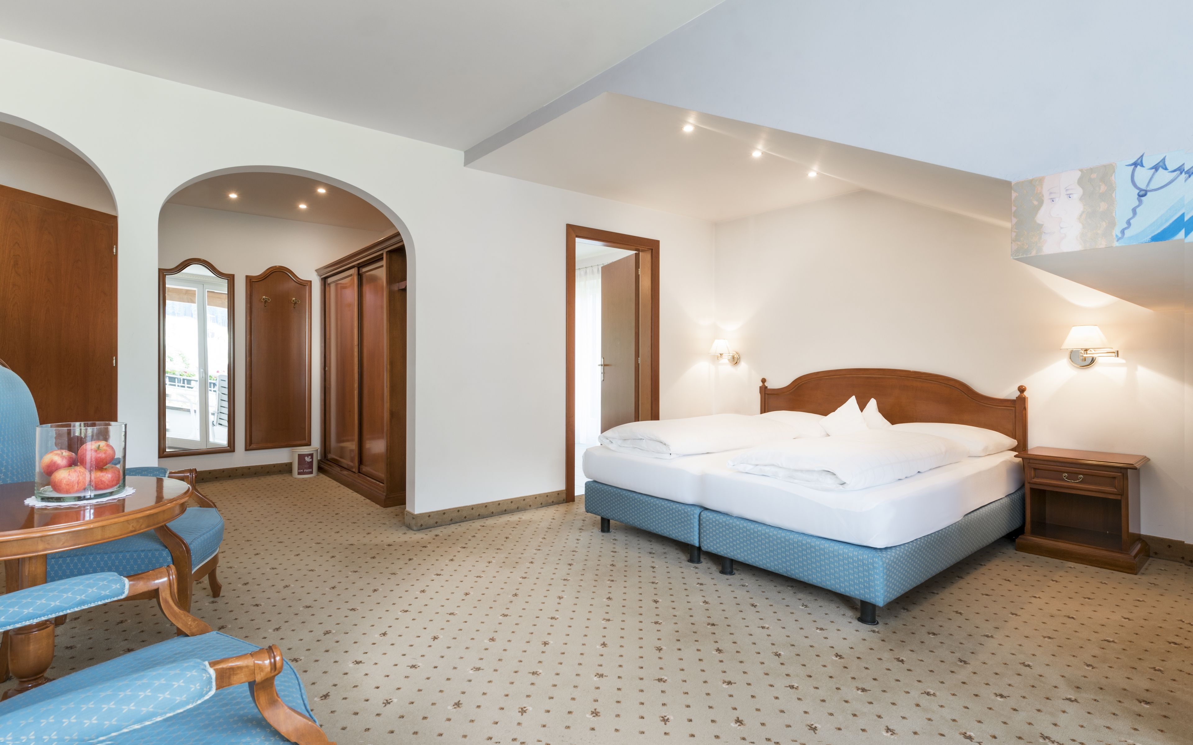 Une chambre d’hôtel avec un grand lit, une armoire en bois, un bureau avec une chaise et une petite table avec des pommes.