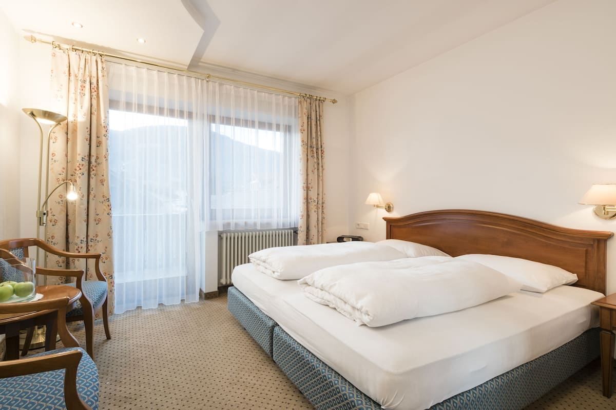 Ein Hotelzimmer mit Doppelbett, zwei Nachttischen, einem Schreibtisch mit Stuhl, einem Heizkörper und einem Fenster mit durchsichtigen Vorhängen.
