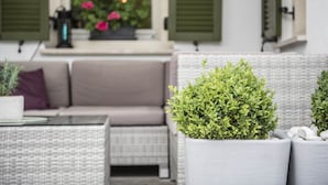 A cozy outdoor seating area with wicker furniture, a potted plant, and a window with shutters. - Boutique Hotel Am Park (Valdaora)