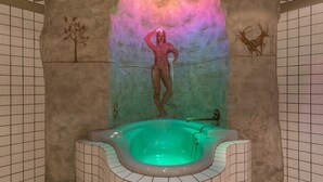 A spa room with a large bathtub, tiled walls, and wall art depicting a tree and a deer. - Boutique Hotel Am Park (Valdaora)