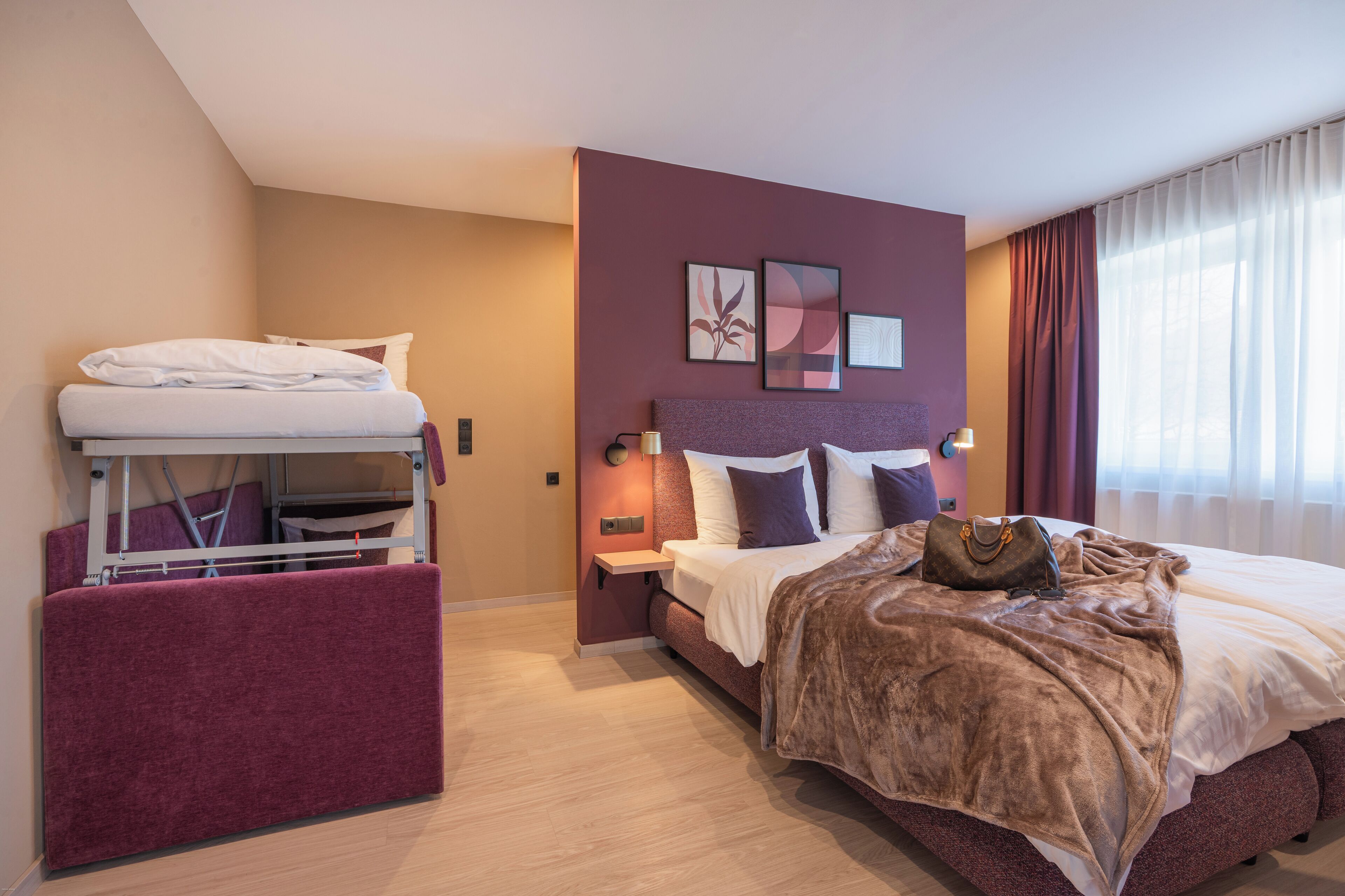 A hotel room with a bunk bed, a large bed, and a view of the outside through sheer curtains.