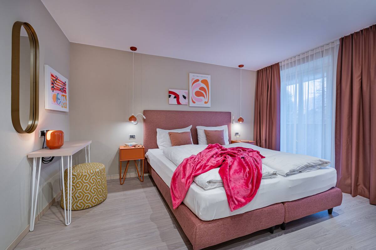 Ein modernes Hotelzimmer mit einem großen Bett, einem Nachttisch, einem kleinen Schreibtisch und einem großen Fenster mit Vorhängen.
