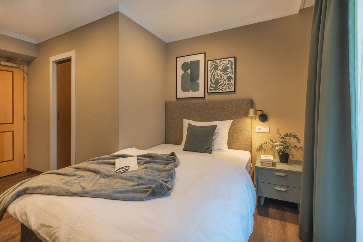 Ein ordentlich gemachtes Bett mit einer grauen Decke, ein Nachttisch mit Lampe und Pflanze sowie zwei gerahmte Kunstwerke an der Wand.