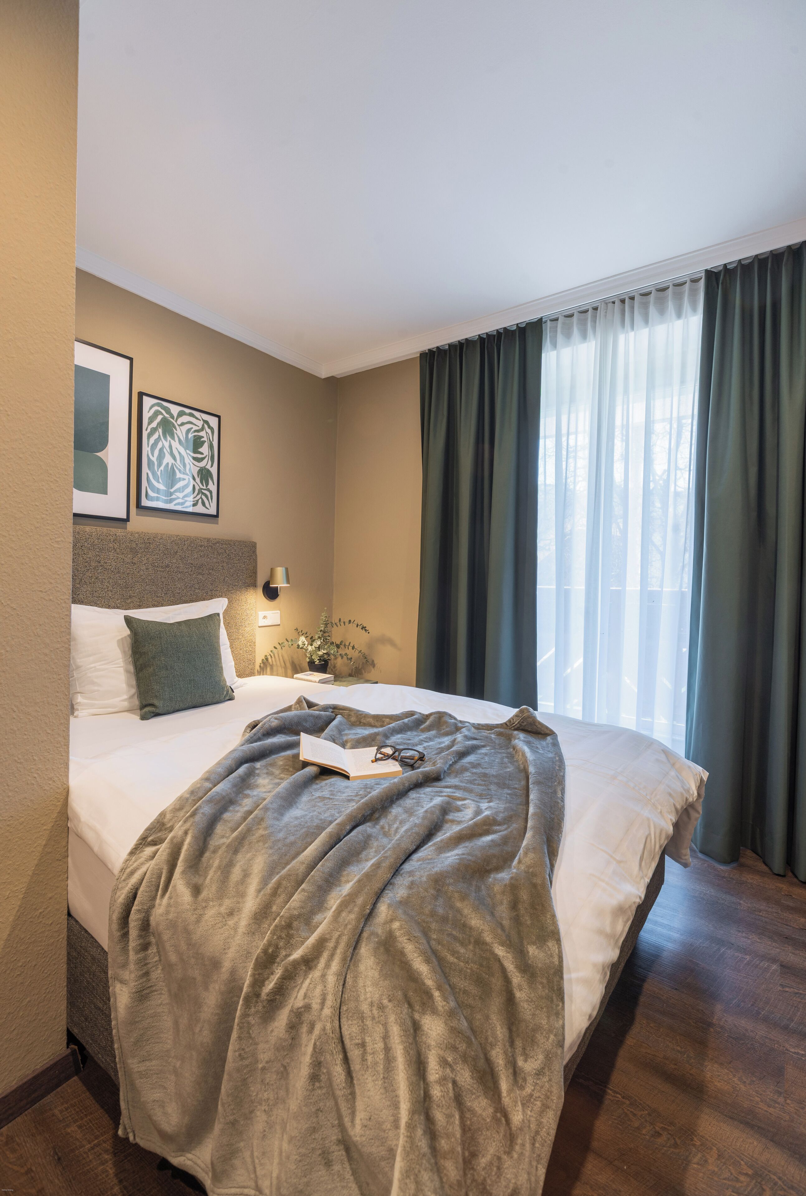 Un lit bien fait, recouvert d’une couverture grise, une lampe de chevet et deux œuvres d’art encadrées accrochées au mur.