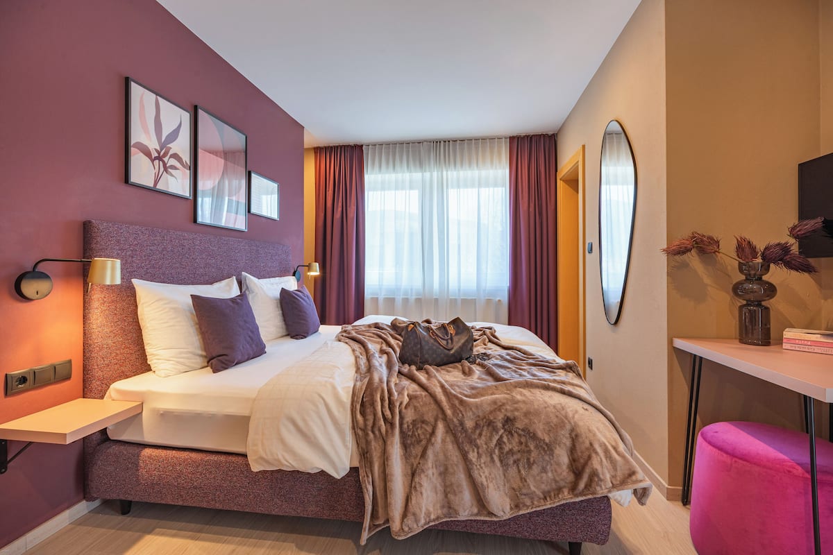 Ein modernes Hotelzimmer mit einem großen Bett, einem Nachttisch, einem Schreibtisch und einem Spiegel.