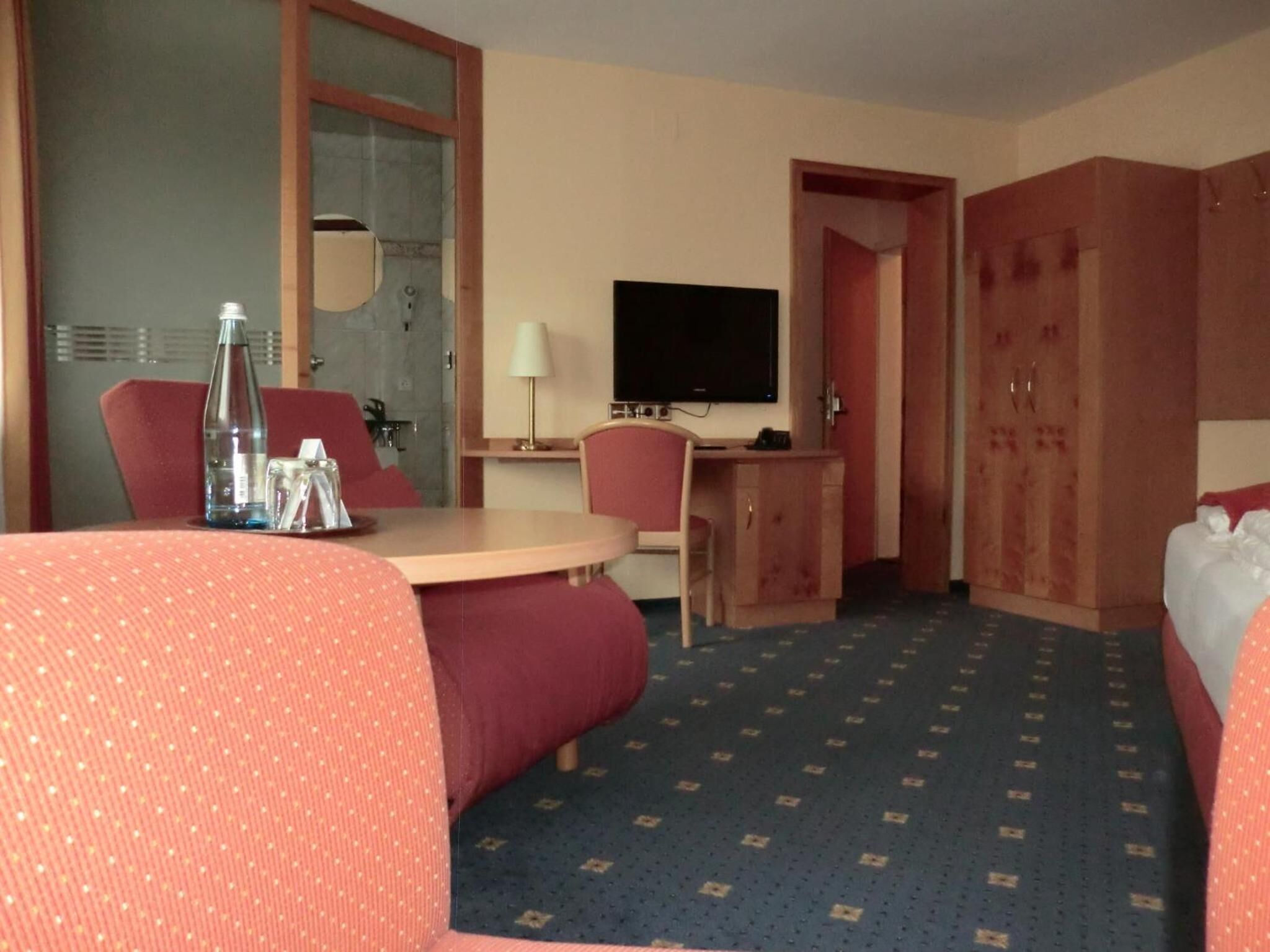 Une chambre d’hôtel comprenant un lit, un bureau avec une télévision, une chaise, une table avec une bouteille et des verres, et une armoire.