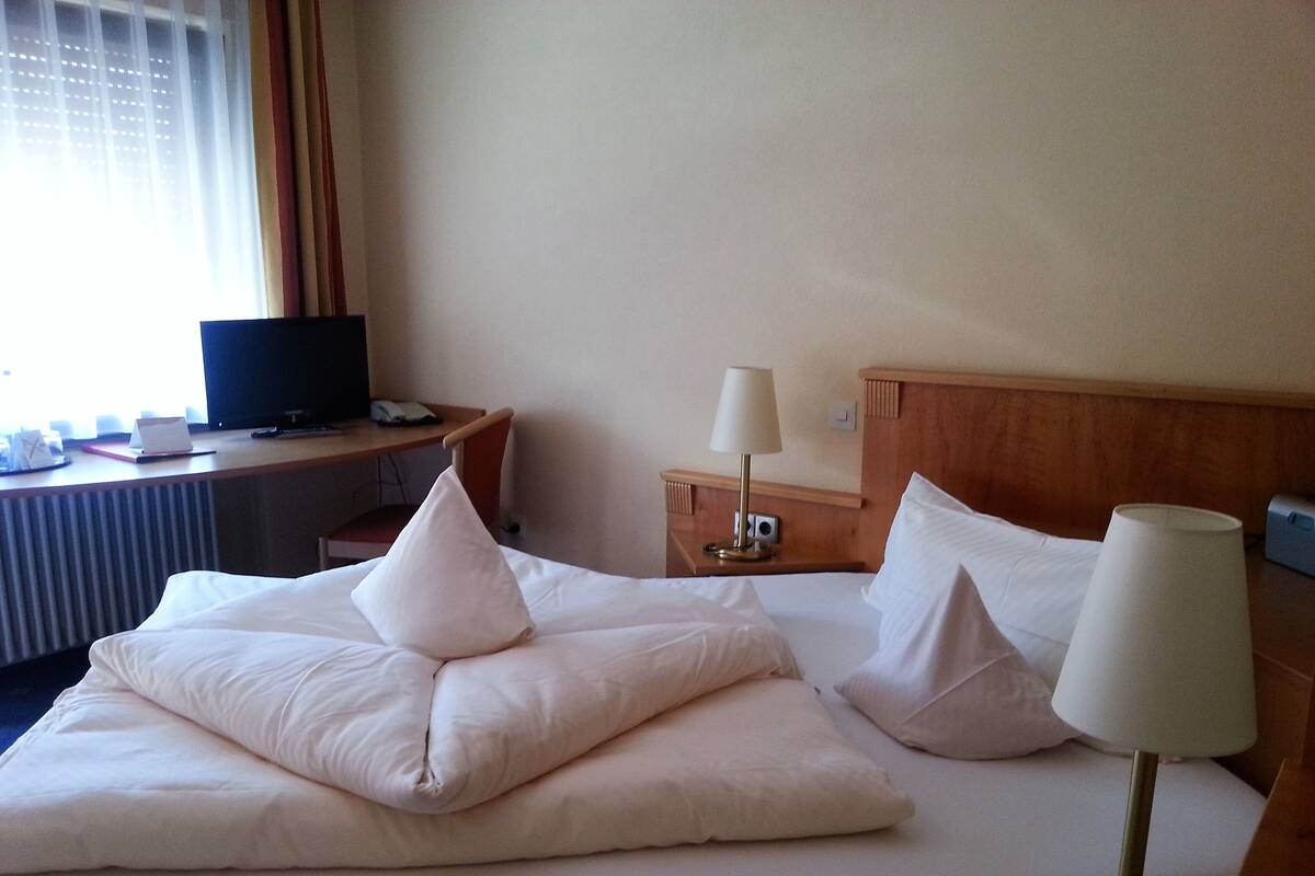 A hotel room with a bed, two pillows, a bedside table with a lamp, a desk with a computer, and a window with curtains.