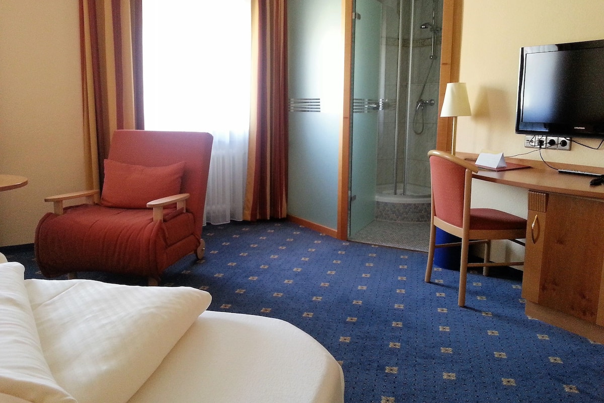 A hotel room with a bed, a desk with a TV, a chair, a shower area, and a window with curtains.
