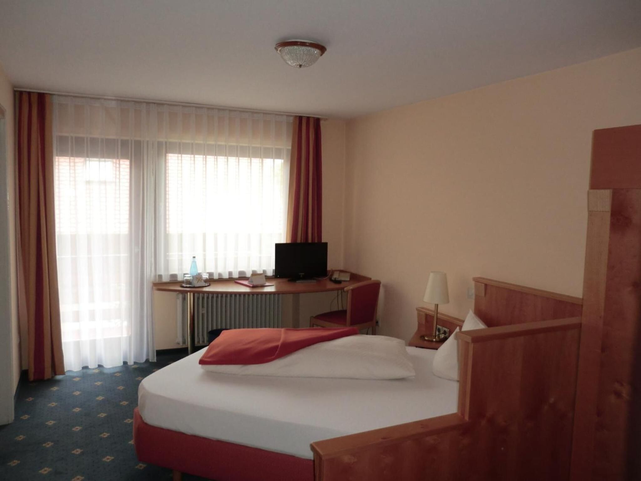 A hotel room with a bed, a desk with a computer, a chair, a lamp, and a window with curtains.