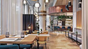 A modern restaurant with wooden tables and chairs, a bar area, and a seating area with a curved wall and plants.