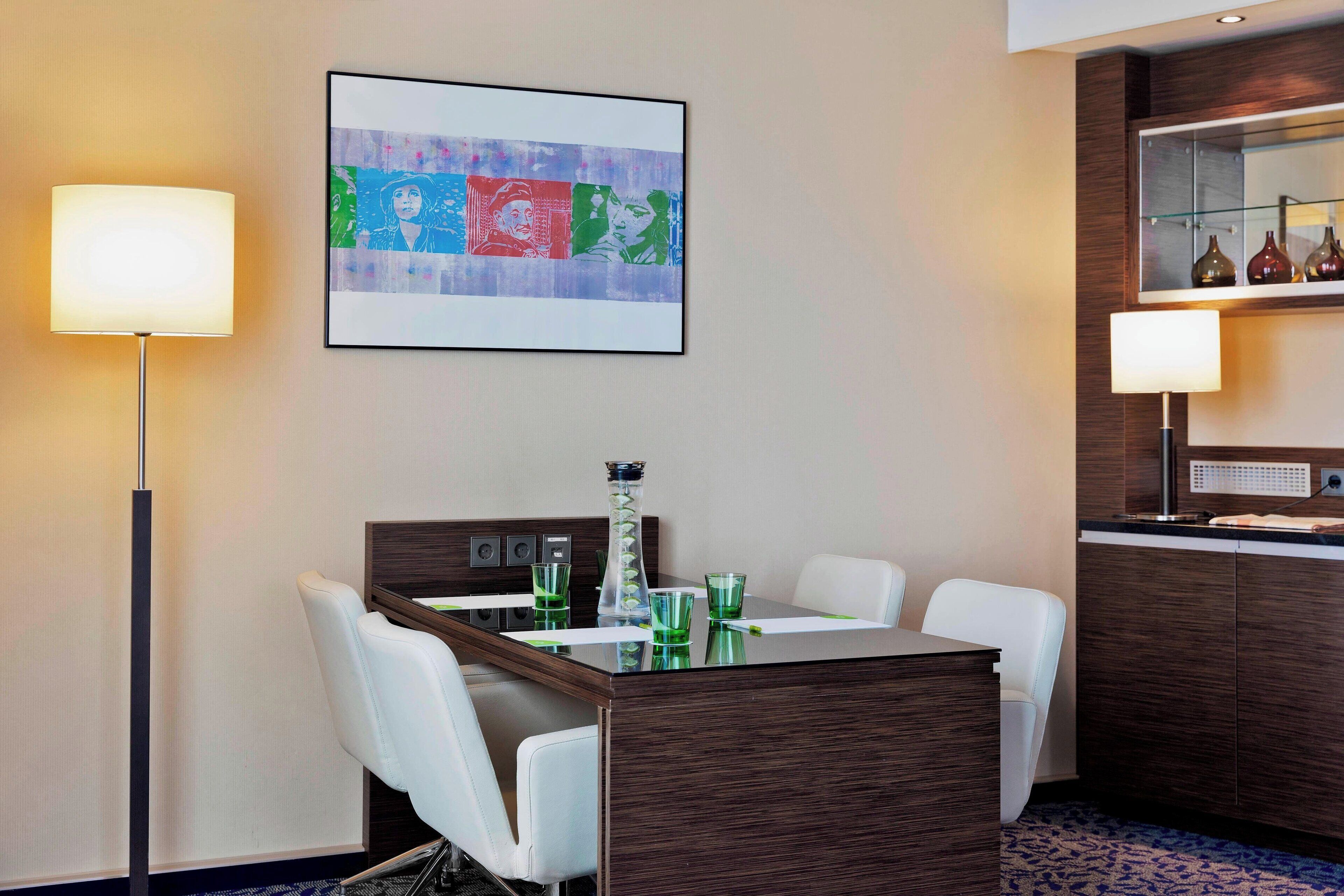 Camera d'albergo con tavolo da pranzo, due sedie, una lampada, un quadro e un minibar.