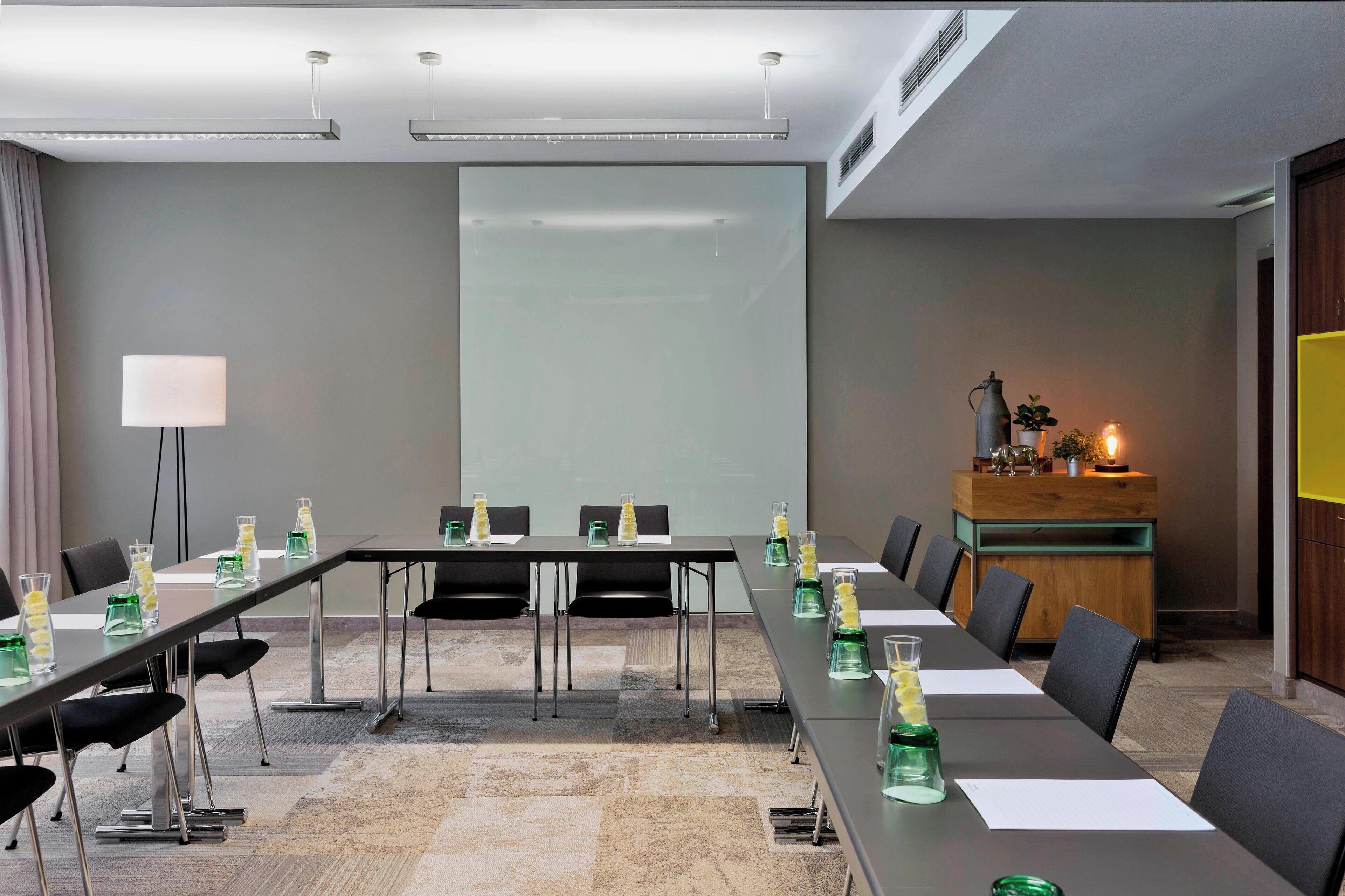 A conference room with long tables, chairs, a projector screen, a desk with a lamp, and a decorative plant.