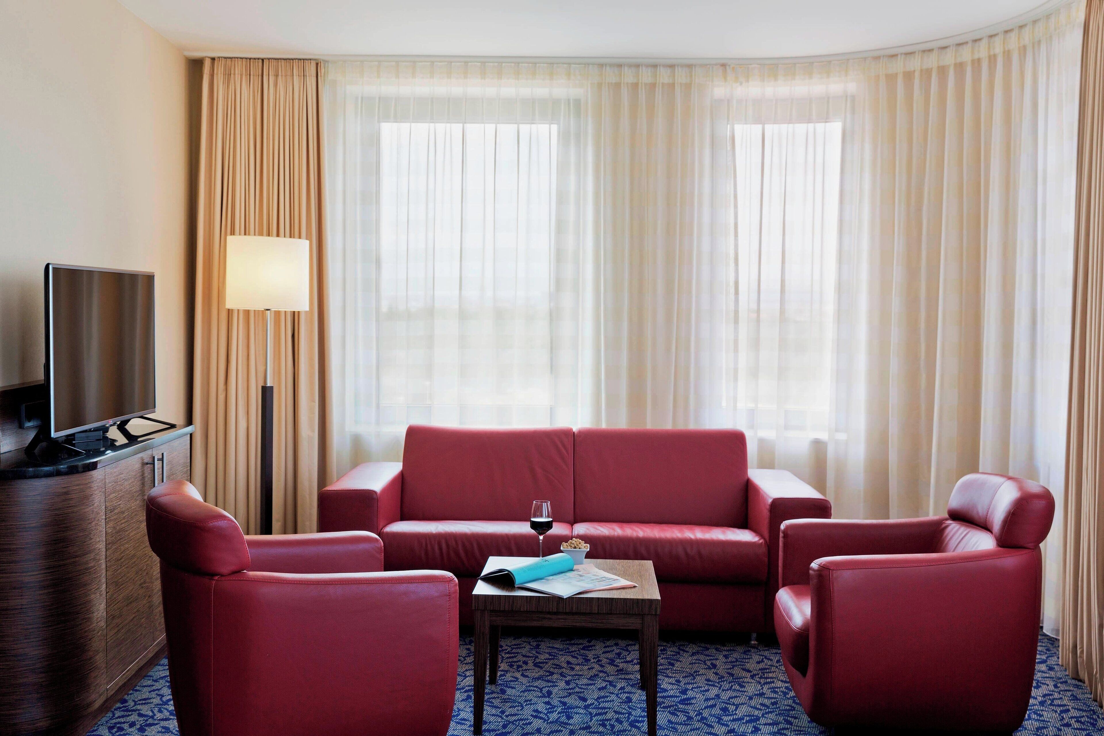 Habitación de hotel con sofá rojo, una mesita con una copa de vino, una lámpara y un televisor sobre un mueble de madera.