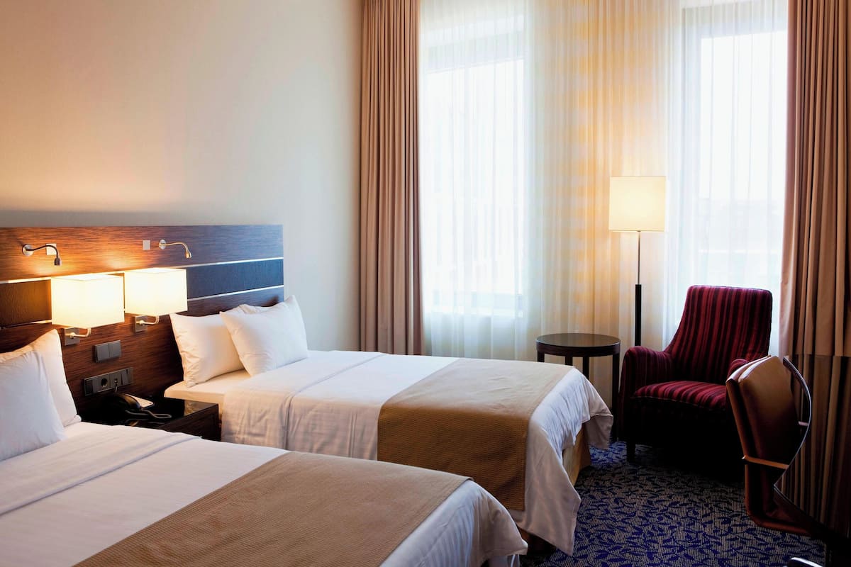 A hotel room with two beds, a desk with a lamp, a chair, and a view of the outdoors through large windows.