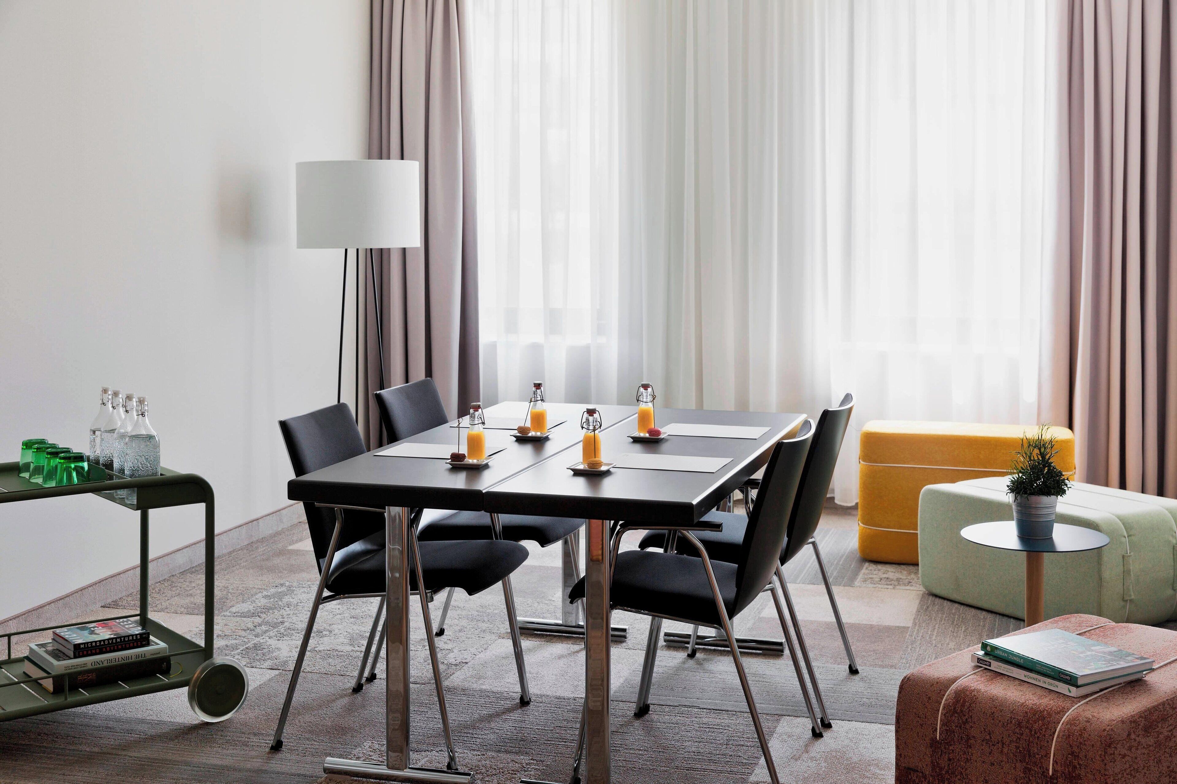 Uma área de jantar moderna com uma mesa posta para seis pessoas, rodeada por cadeiras pretas. Uma pequena mesa redonda com uma luminária e uma planta em vaso é visível.