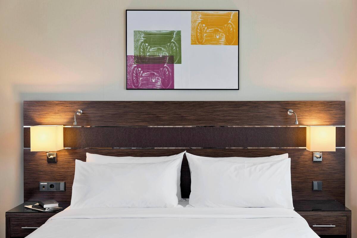 A hotel room with a wooden headboard, two bedside lamps, a bed with white linens, and a framed artwork above the bed.