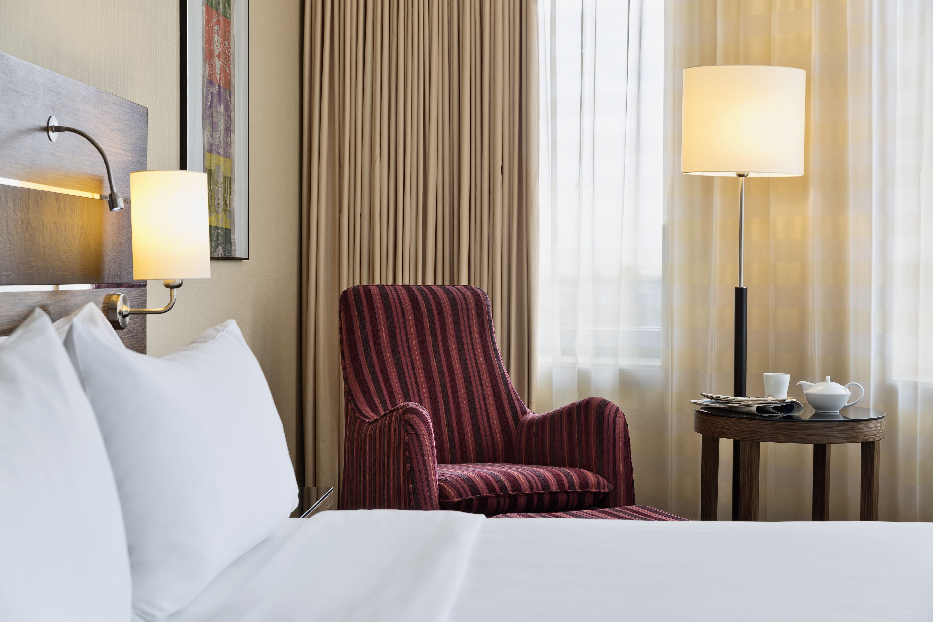 A hotel room with a bed, a striped armchair, a small round table with a lamp, and a painting on the wall.