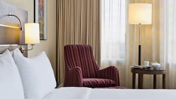 A hotel room with a bed, a striped armchair, a small round table with a lamp, and a painting on the wall.