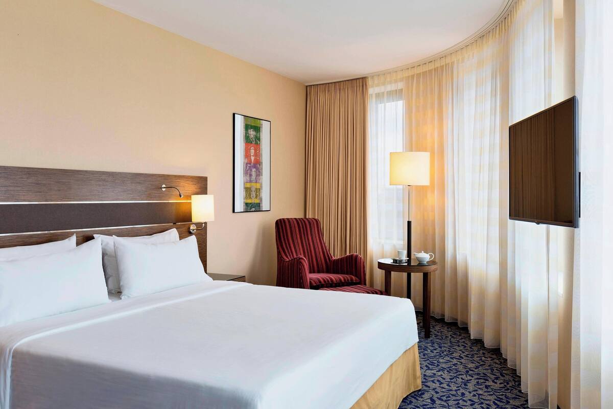 A hotel room with a large bed, a red armchair, a small table with a lamp, a television, and a painting on the wall.