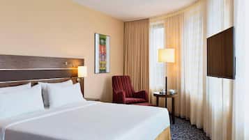 A hotel room with a large bed, a red armchair, a small table with a lamp, a television, and a painting on the wall.