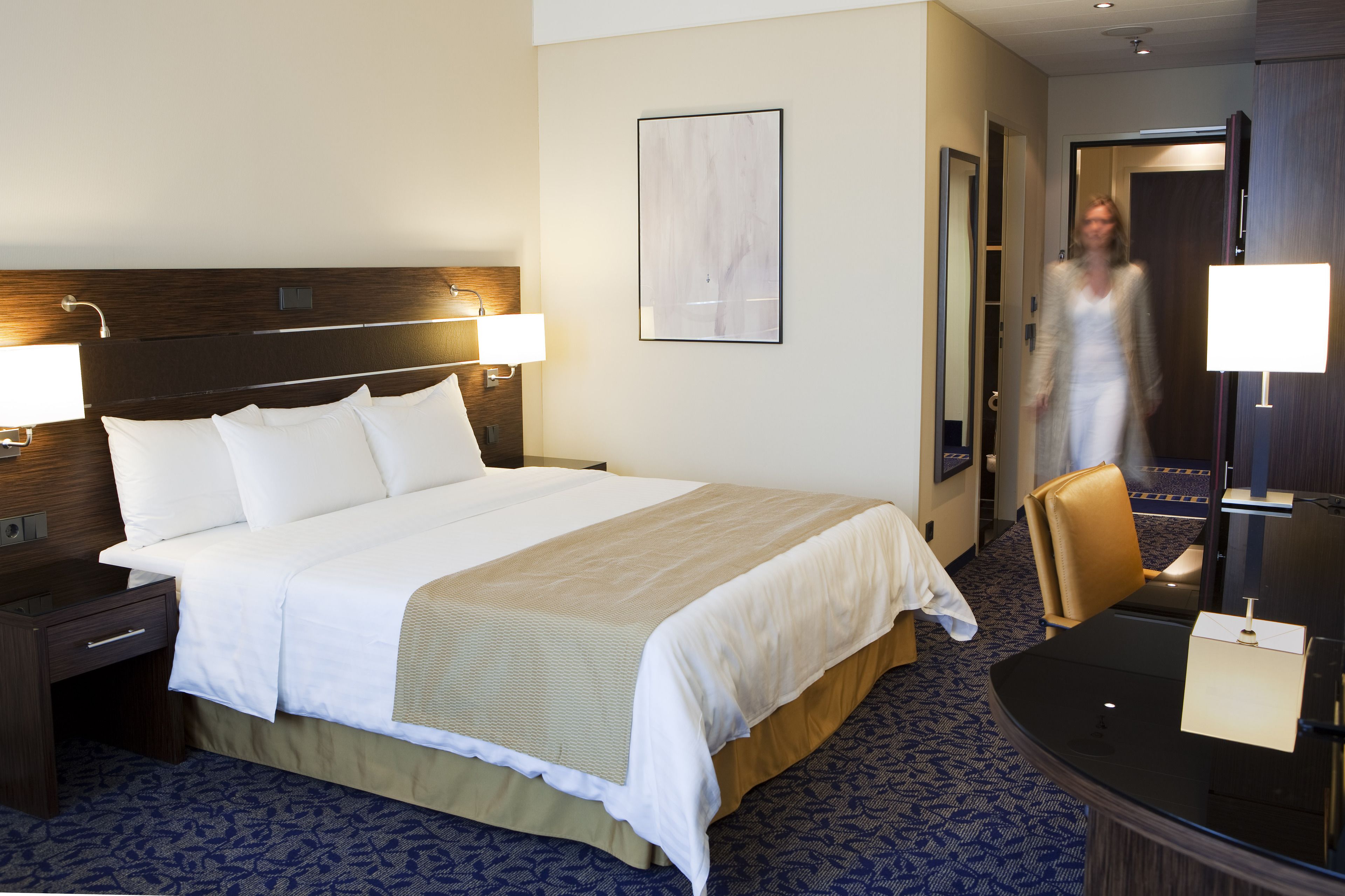 A hotel room with a large bed, a desk, a chair, and a mirror.