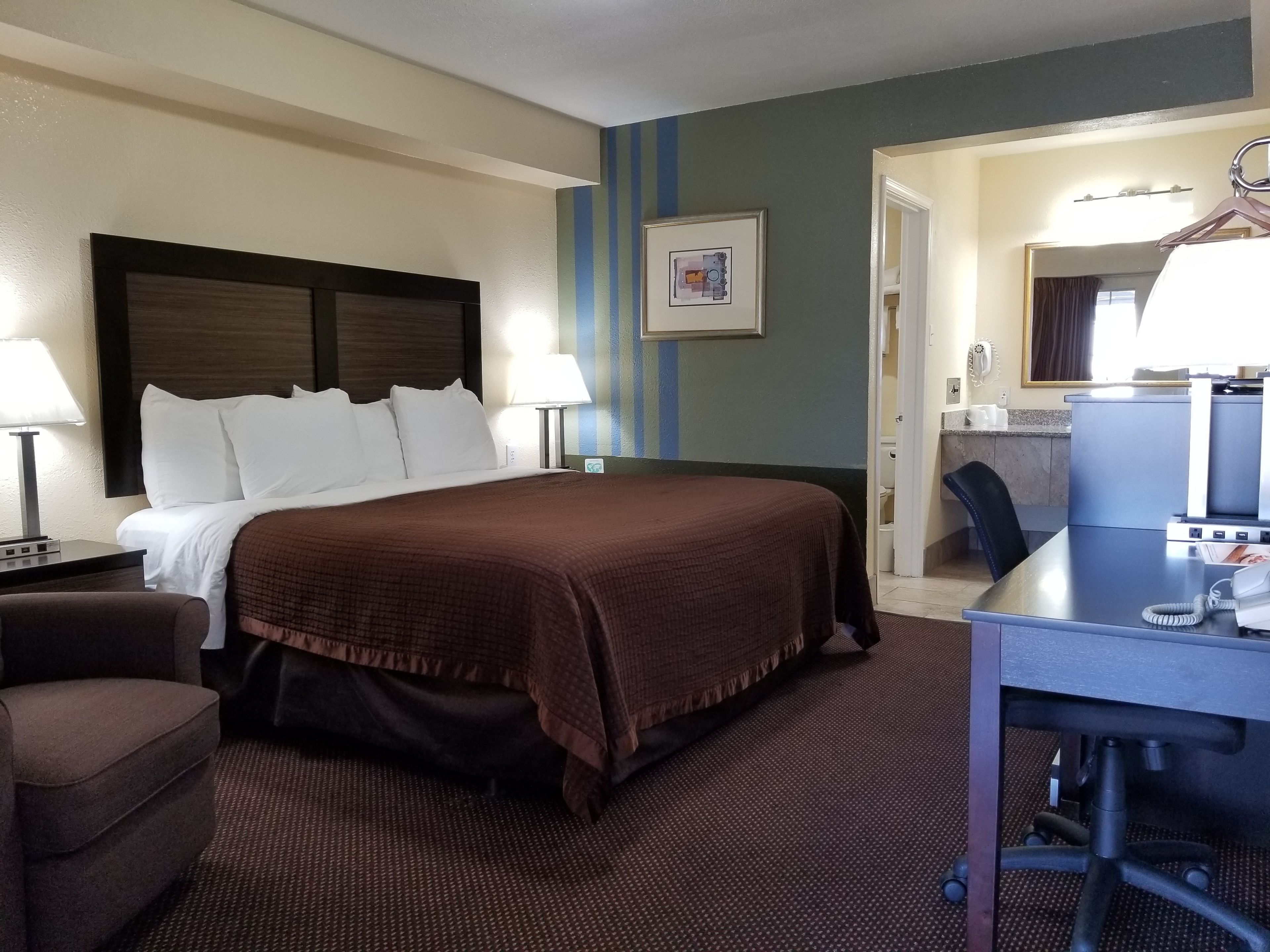Ein Hotelzimmer mit einem großen Bett, einem Schreibtisch, einem Sessel, einer Lampe, einem gerahmten Bild an der Wand und einem durchgehenden Türspalt einsehbaren Badezimmer.