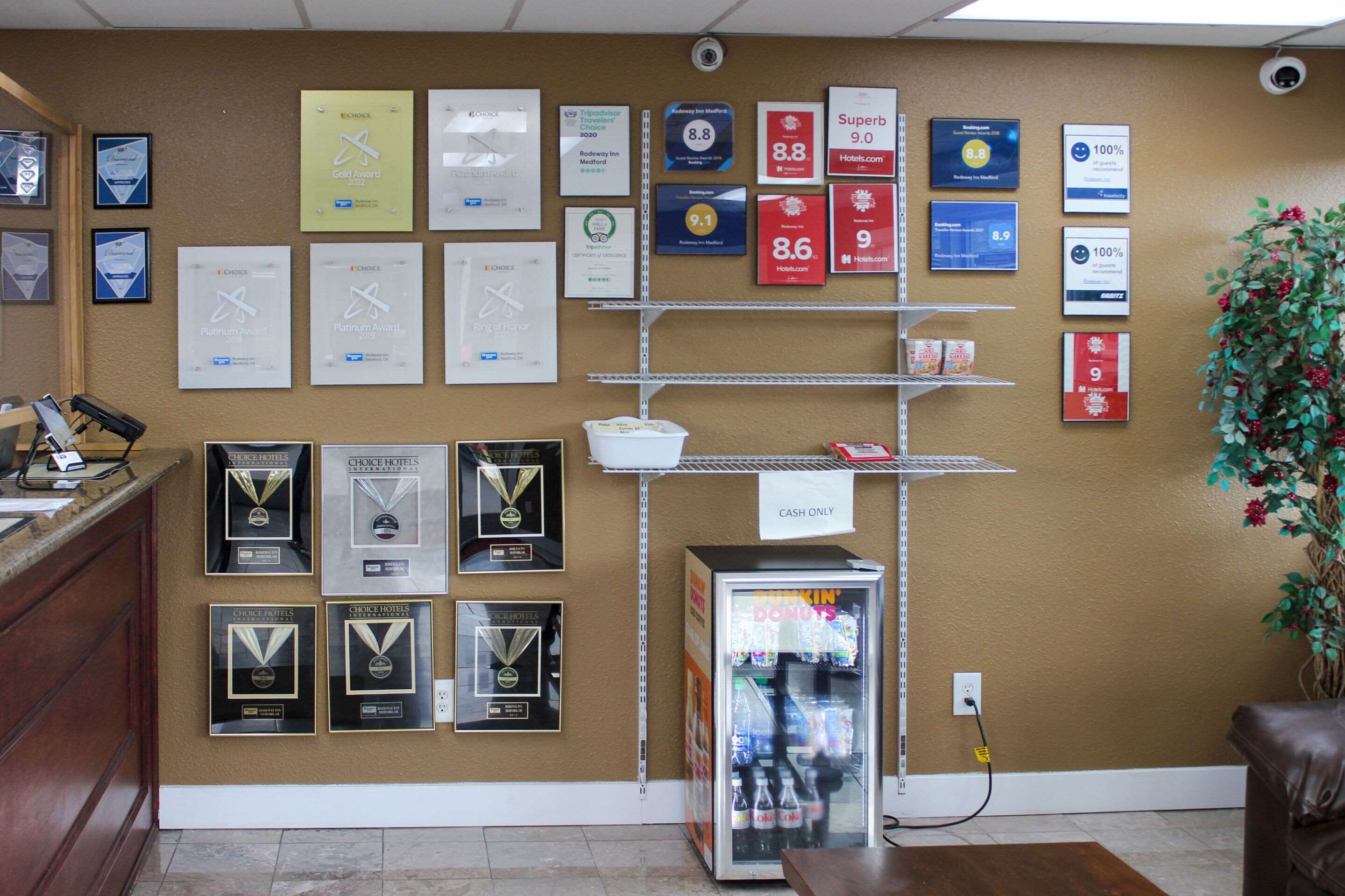 A hotel reception area with framed awards, a cash-only sign, a Dunkin' Donuts refrigerator, and a potted plant.
