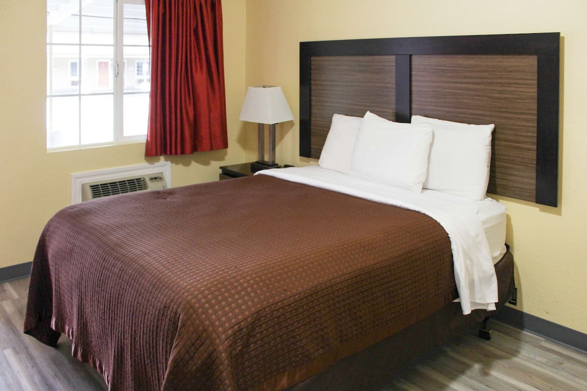 Quarto de hotel com uma cama grande, mesa de cabeceira, luminária, janela com cortinas e ar condicionado.
