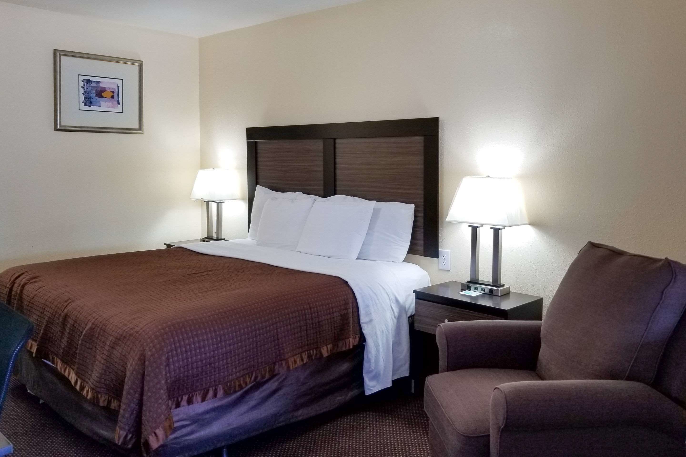 A hotel room with a large bed, two bedside lamps, a brown chair, and a framed picture on the wall.