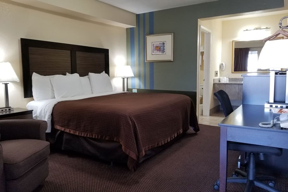 Quarto de hotel com uma cama grande, uma mesa, uma cadeira, uma luminária, um quadro na parede e um banheiro visível através de uma porta aberta.