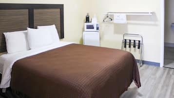 A hotel room with a bed, white pillows, a brown blanket, a white microwave, a small refrigerator, and a towel rack with hangers.