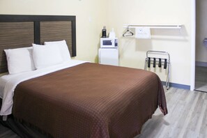 A hotel room with a bed, white pillows, a brown blanket, a white microwave, a small refrigerator, and a towel rack with hangers.