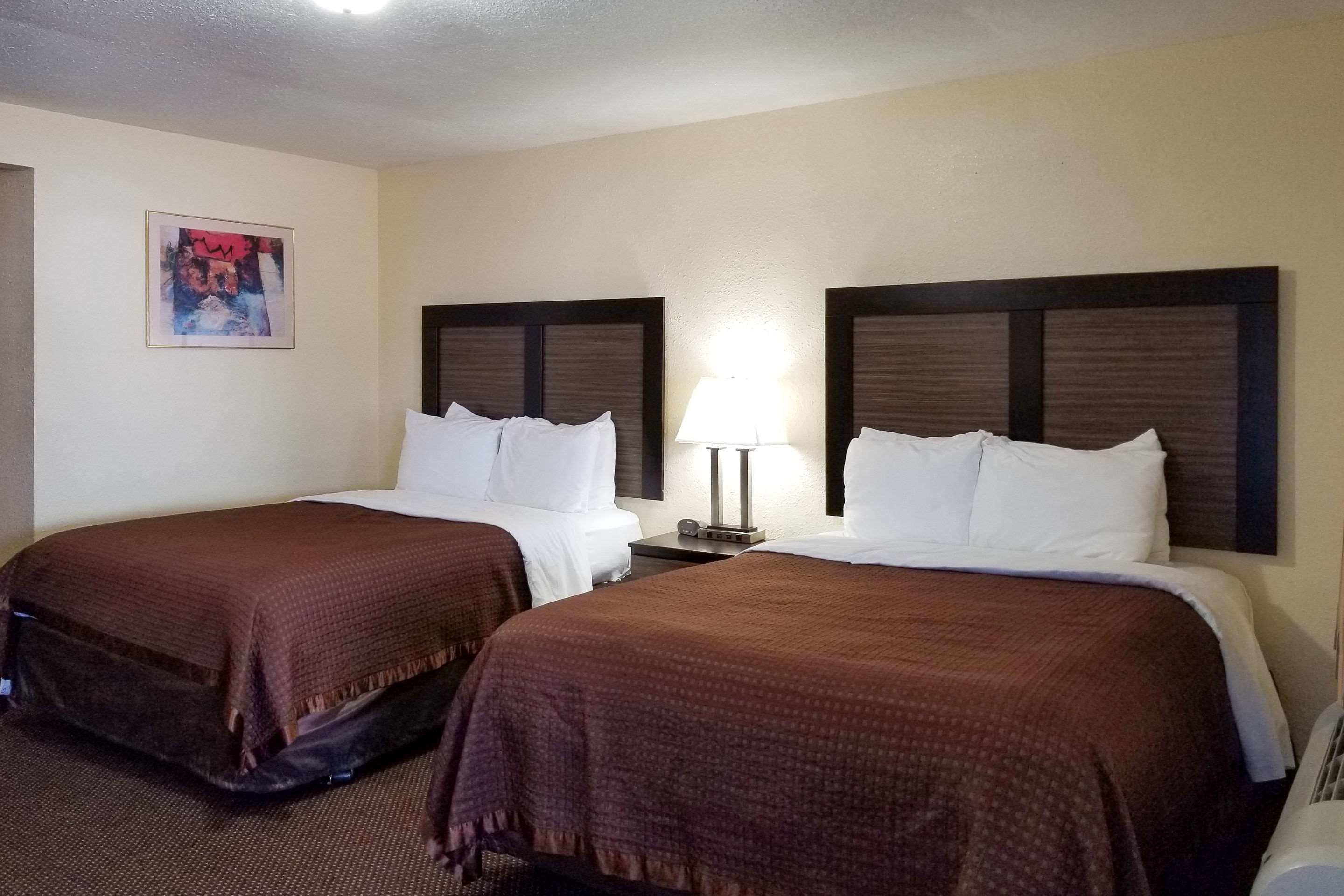 A hotel room with two beds, each with a brown blanket, a bedside table with a lamp, and a framed picture on the wall.
