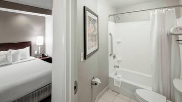 A hotel room with a bed, a bedside table with a lamp, a framed picture on the wall, and a bathroom with a bathtub, toilet, and shower curtain.