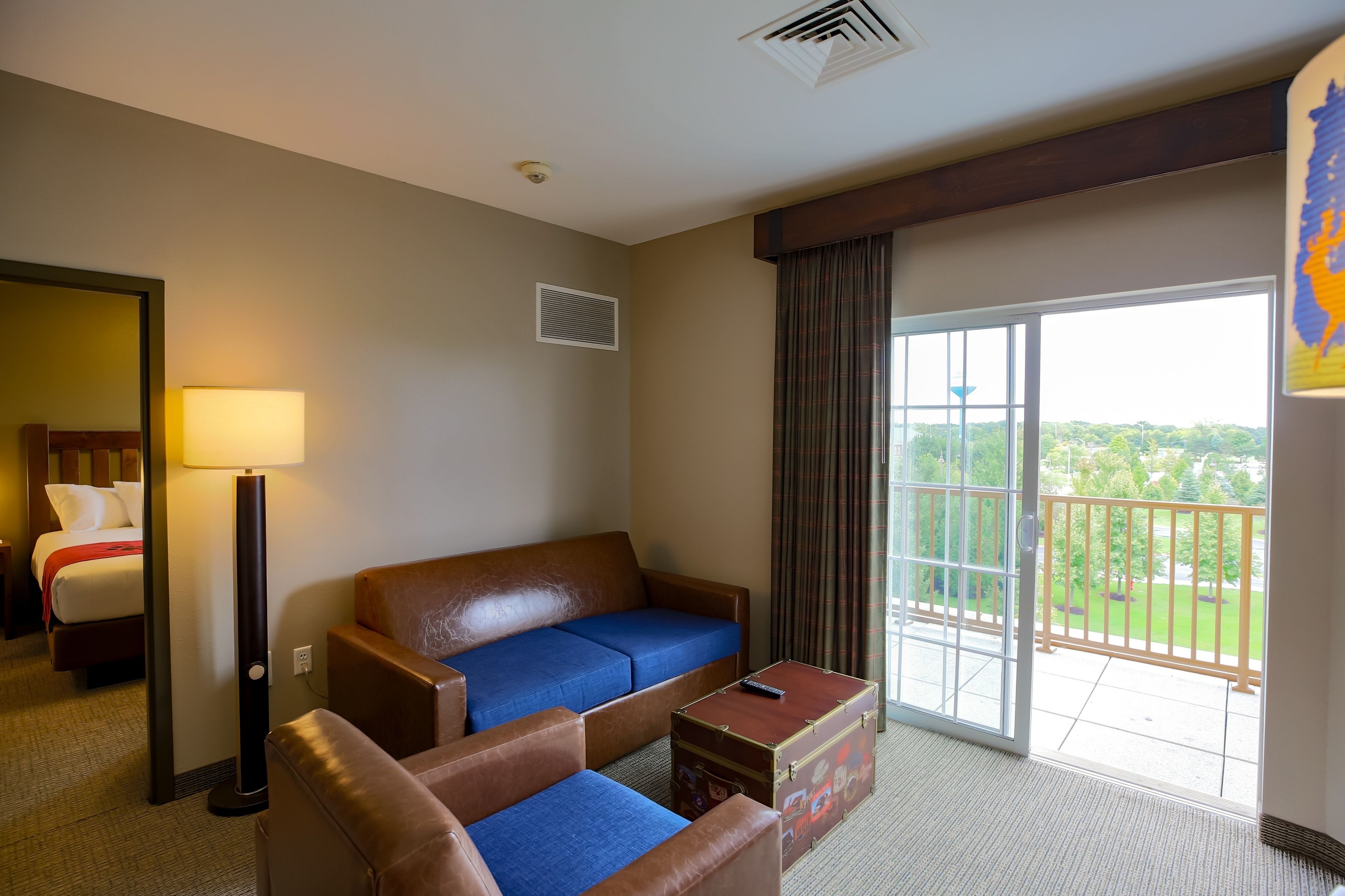 Habitación de hotel con sofá, dos sillones, una mesita, una lámpara, puerta corrediza de vidrio a un balcón y una cama con una colcha roja.