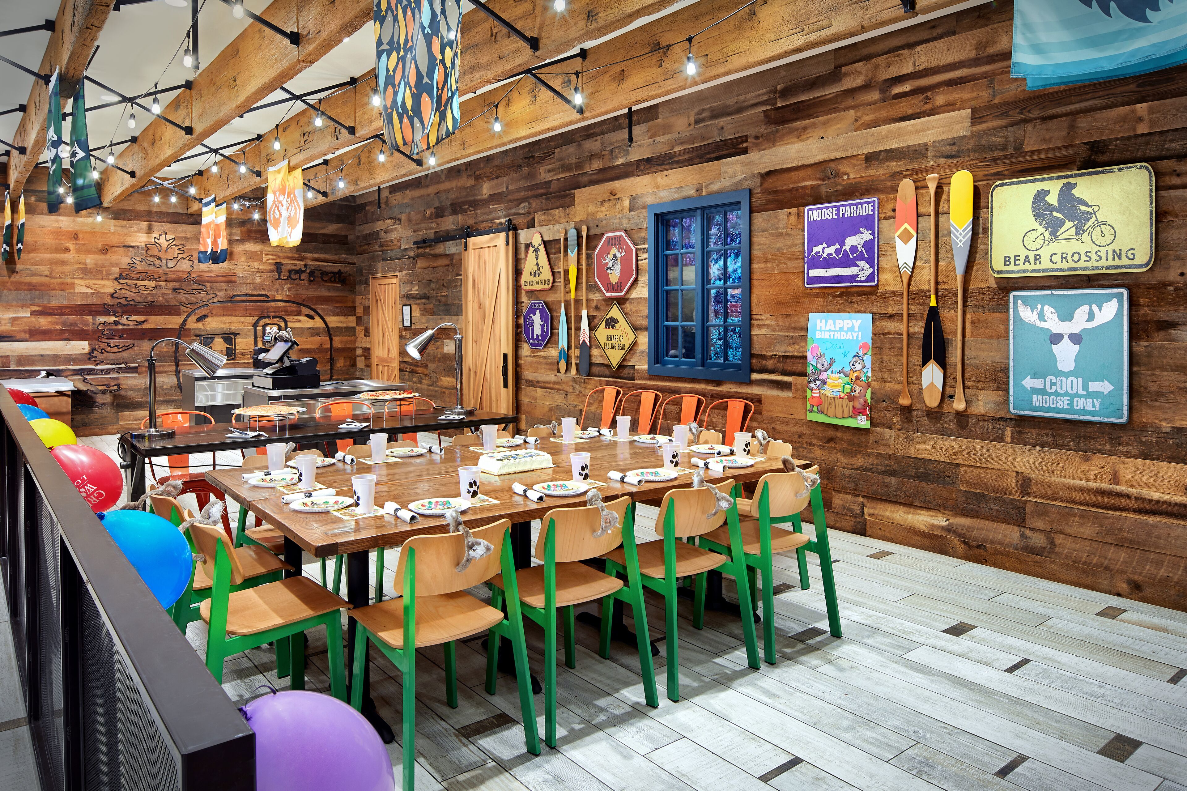 A rustic wooden interior with a long dining table set for a meal, surrounded by colorful chairs.