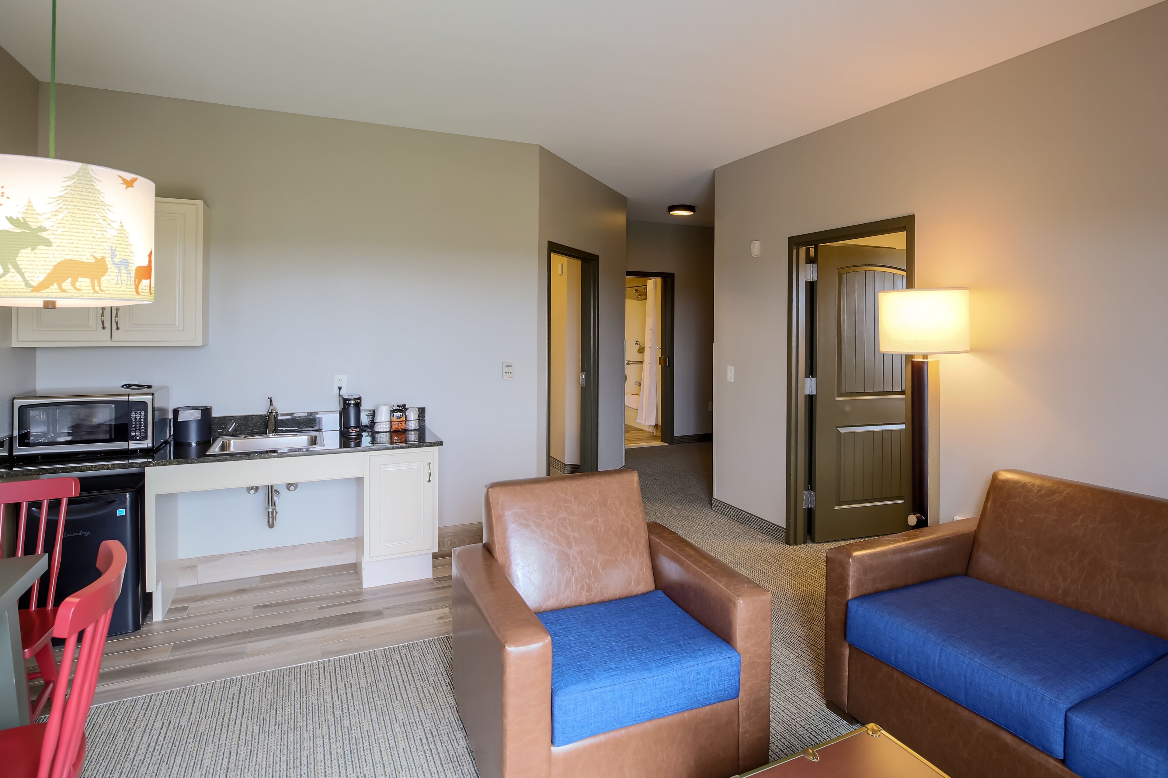 A compact hotel room with a kitchenette, a sofa, a chair, and a lamp.