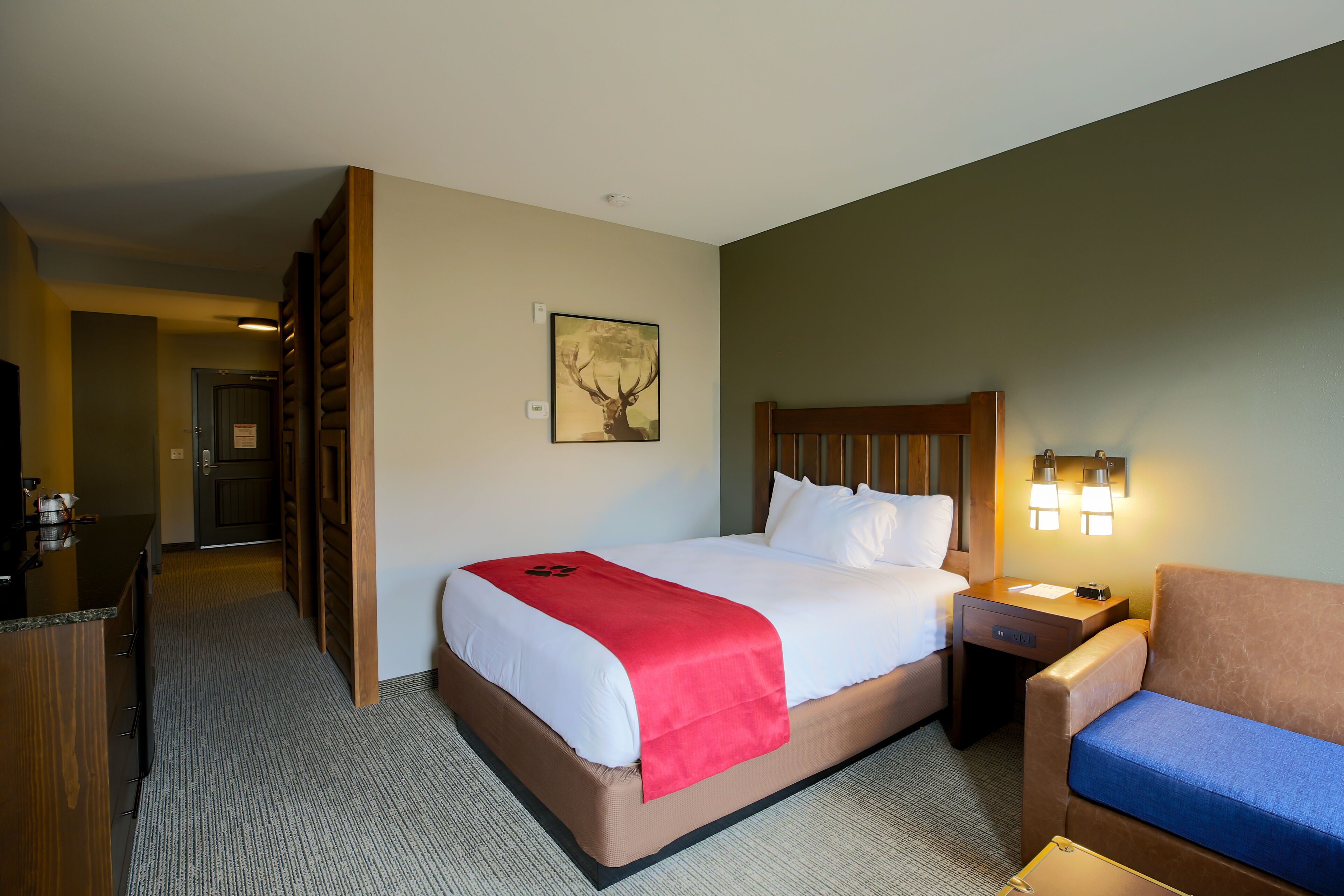 A hotel room with a large bed, a wooden headboard, a nightstand with a lamp, a brown leather sofa, and a desk with a chair.