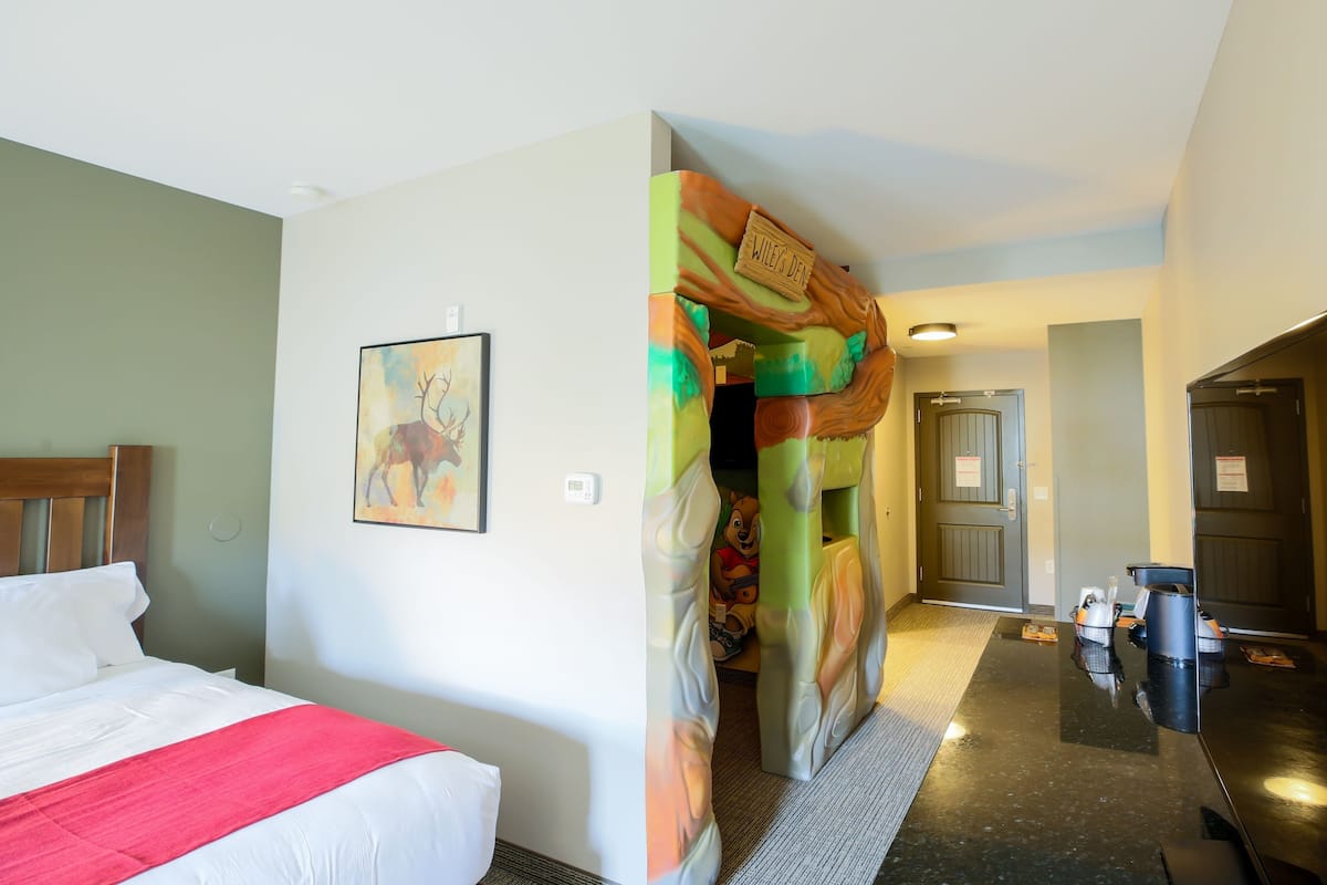 A hotel room with a bed, a painting, a colorful tree-themed door, and a kitchenette area.