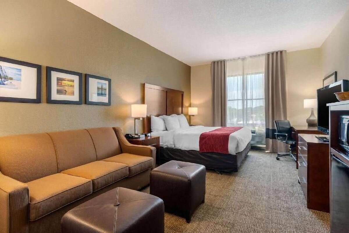 Ein Hotelzimmer mit Bett, Sofa, Hocker, Schreibtisch, Fernseher und gerahmten Bildern an der Wand.