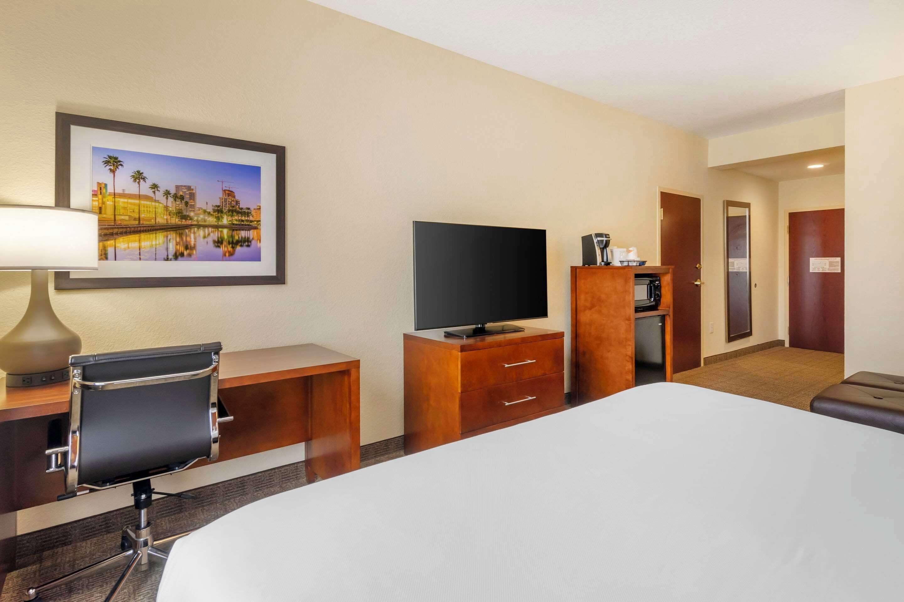 A hotel room with a bed, a desk with a chair, a television, a coffee machine, and a framed picture on the wall.