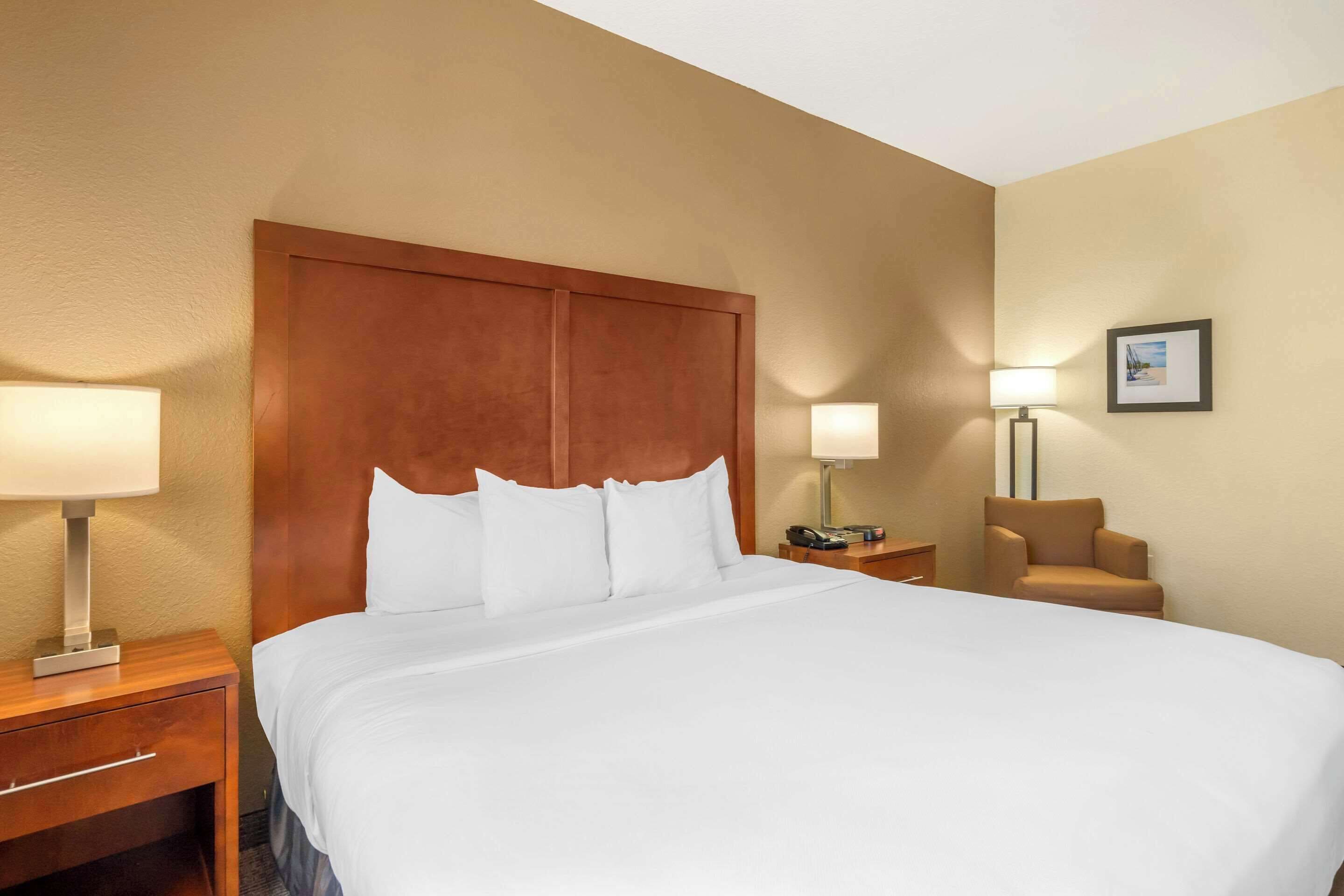 A hotel room with a large bed, wooden headboard, two bedside tables with lamps, a chair, and a framed picture on the wall.