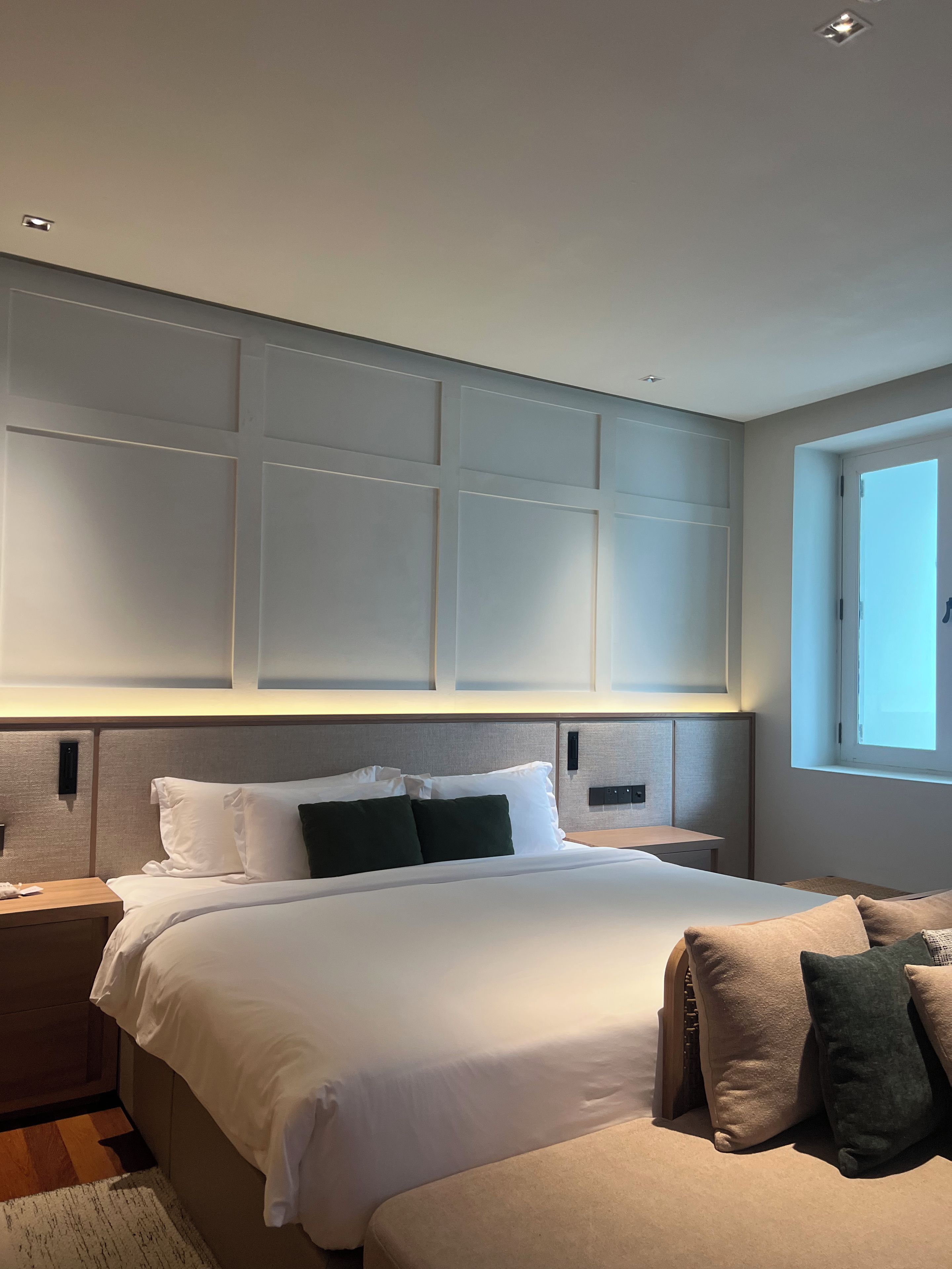 A hotel room with a large bed, bedside tables, a nightstand, a window, and a wall panel design.