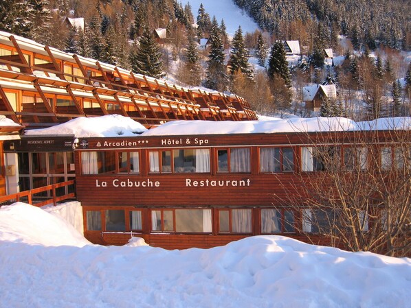 Un edificio de madera con un letrero que dice "La Cabuche Restaurant" y "Arcaden Hotel & Spa" en una zona montañosa nevada.
